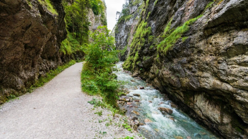 Kundler Klamm in Kundl bei Wörgl Tirol Gepflegter Kiesweg führt durch eine enge, grüne Schlucht. Ein Bach verläuft parallel zum Weg, flankiert von hoch aufragenden Felswänden und üppiger Vegetation.