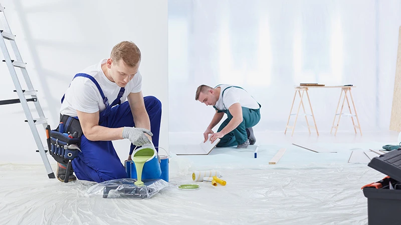Zwei Maler aus einer professionellen Malerfirma am arbeiten. Maler füllt Farbe aus einem Farbeimer um. Hinter ihm arbeitet ein anderer Malermeister, und verlegt einen Schutz für den Boden als Vorbereitung.
