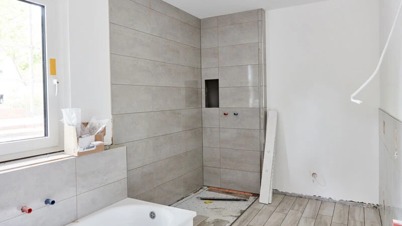 Unfertiges Badezimmer mit grauen Fliesen und vorbereiteter Dusche, Renovierungsarbeiten.