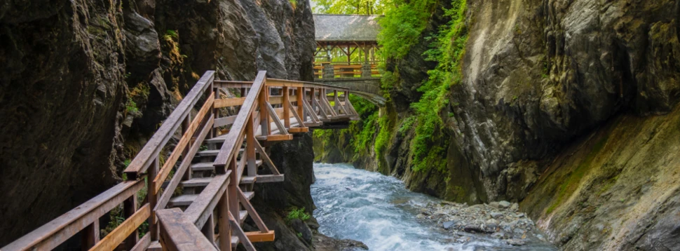 Holzsteg schlängelt sich entlang einer engen, felsigen Schlucht über einem rauschenden Fluss umgeben von steilen Felswänden und grünem Laubwald im Hintergrund