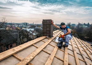 Dachsanierung Wolfsberg Dachdecker hockt auf ungedecktem Dach unter Schornstein und beginnt mit der Eindeckung der Dachziegel. Himmel im Hintergrund blau und kleine Wolken.