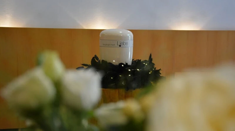 Sanftes Licht, weiße Rosen und die Aufschrift „Vergänglichkeit – Liebe" auf der Urne verbinden Trauer und Erinnerung zu einem stillen Moment des Gedenkens.