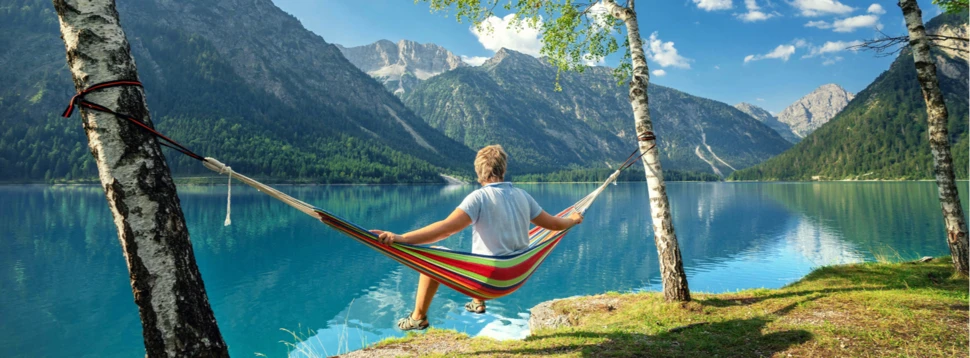 Mann in Hängematte zwischen zwei Bäumen an einem blauen See, im Hintergrund Bäume und Wälder