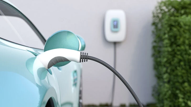 Elektroauto-Ladestation mit eingestecktem Ladekabel an einem türkisfarbenen Elektrofahrzeug, im Hintergrund eine Wallbox und grüne Hecke, Nahaufnahme der Ladebuchse