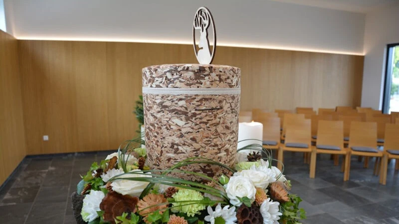 Eine Urne aus natürlichen Materialien, umgeben von einem liebevoll gestalteten Blumenkranz, schafft in der modernen Trauerhalle einen würdevollen Rahmen für den letzten Abschied.