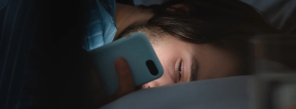 Ein junger Mann liegt schlaflos im Bett und starrt auf sein Handy. Er ist offenbar süchtig nach Social Media. Digital Detox würde ihm guttun.