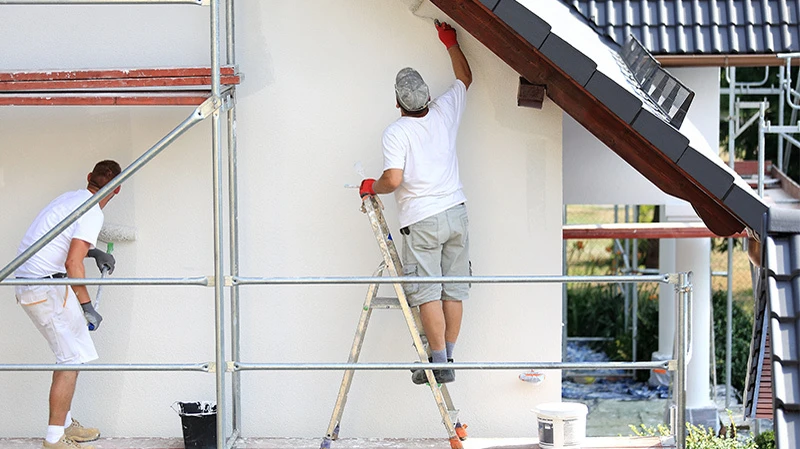 Zwei Maler aus einem professionellen Malerbetrieb streichen ein Einfamilienhaus in Wels. Professioneller Malerbetrieb am Werk. Zwei Maler stehen auf einem Gerüst und streichen mit einer weißen Farbrolle die Fassade eines Einfamilienhaus in Wels.